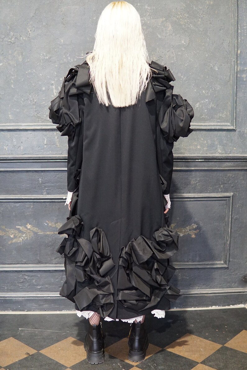 Puede incluir: Un vestido negro con falda larga y grandes volantes. El vestido tiene un corpi&ntilde;o ajustado y mangas largas. La modelo lleva botas negras.