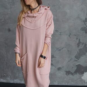 May include: A pink hooded sweatshirt dress with frayed edges and two large pockets. The dress is worn by a person wearing a brown fedora hat.
