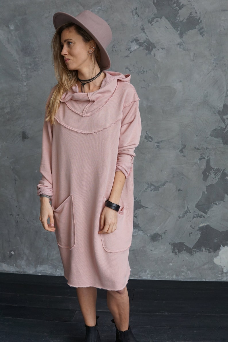 May include: A pink hooded sweatshirt dress with frayed edges and two large pockets. The dress is worn by a person wearing a brown fedora hat.