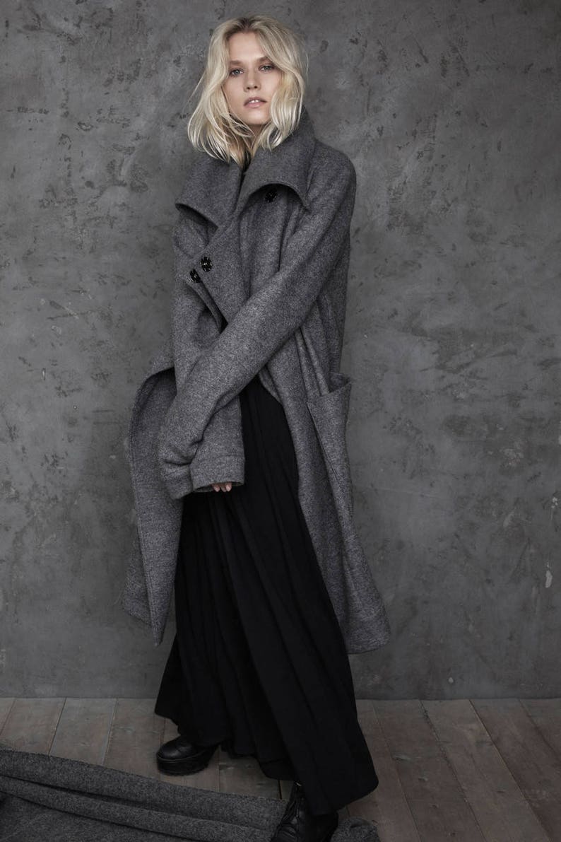 Pu&ograve; includere: Una donna indossa un cappotto grigio a maniche lunghe con un ampio colletto e una gonna maxi nera. &Egrave; in piedi davanti a un muro grigio con una texture.