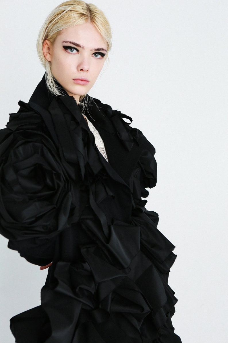 Puede incluir: Una mujer con un vestido negro con grandes mangas con volantes. El vestido est&aacute; hecho de una tela suave y fluida y tiene una silueta voluminosa y dram&aacute;tica.