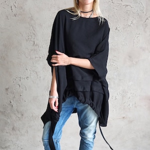 Può includere: Una donna indossa una tunica nera oversize con un dettaglio arricciato in vita, jeans blu, un cappello fedora grigio e occhiali da sole neri.