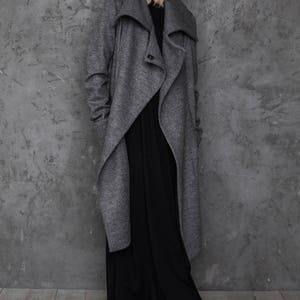Pu&ograve; includere: Una donna indossa un lungo cappotto avvolgente grigio con un ampio colletto e un abito maxi nero. &Egrave; in piedi davanti a un muro di cemento grigio.