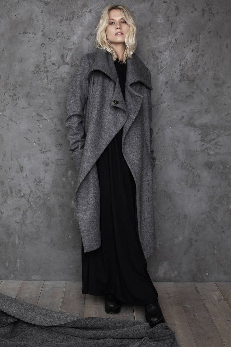 Pu&ograve; includere: Una donna indossa un lungo cappotto avvolgente grigio con un ampio colletto e un abito maxi nero. &Egrave; in piedi davanti a un muro di cemento grigio.