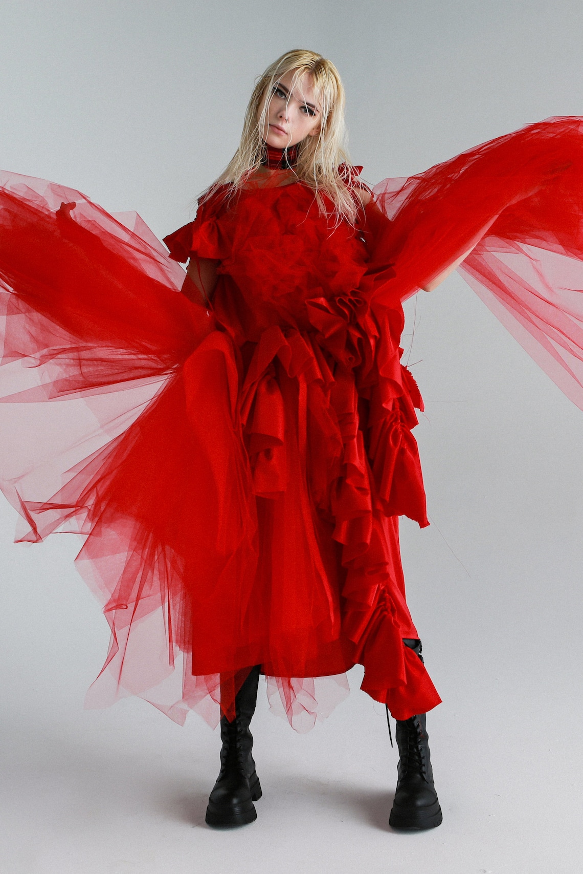 May include: A woman wearing a red tulle dress with ruffles and black boots. The dress has a flowing skirt and the woman is posing with her arms outstretched.