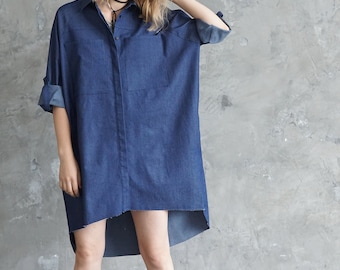 May include: A woman wearing a long, dark blue denim shirt dress with a high-low hemline. She is standing in front of a gray concrete wall and wearing black ankle boots.