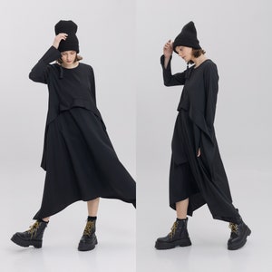 Puede incluir: Dos modelos visten vestidos negros de manga larga con dobladillos asimétricos. Ambos modelos llevan gorros negros y botas de cordones negras.