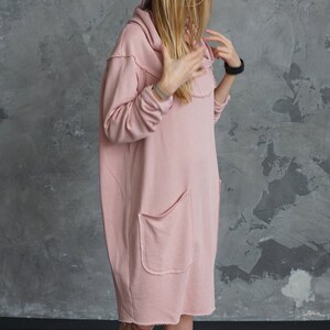 May include: A pink oversized hooded sweatshirt dress with a single front pocket and raw edges. The dress is worn by a person wearing a brown fedora hat and black ankle boots.