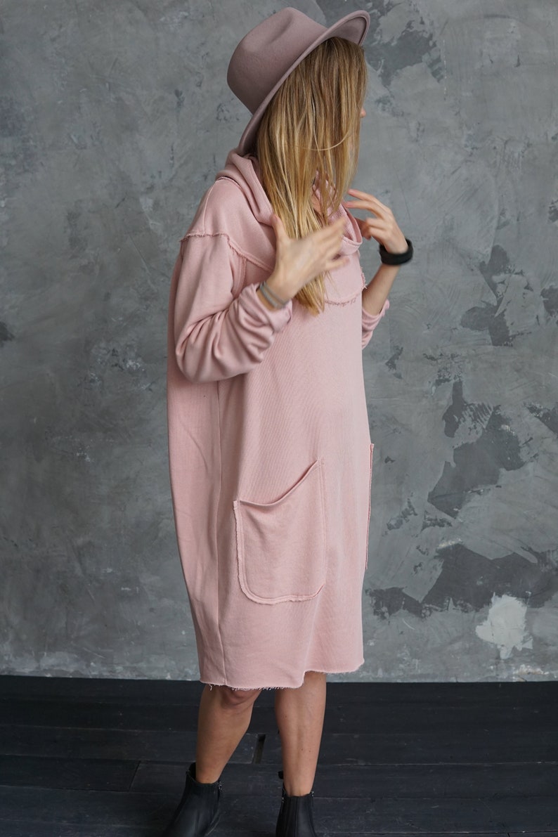 May include: A pink oversized hooded sweatshirt dress with a single front pocket and raw edges. The dress is worn by a person wearing a brown fedora hat and black ankle boots.