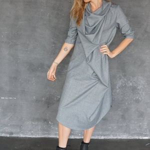 Puede incluir: Un vestido gris de cuello de pico con manga larga y corte holgado. El vestido se combina con botines negros de tobillo y un fedora negro.