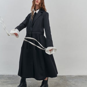 May include: A woman wearing a black long-sleeved coat with a white collar and a black tie. She is wearing black boots and holding a white ribbon.
