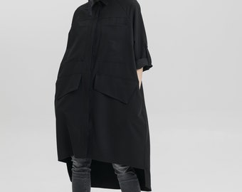 May include: A black long-sleeved shirt with a high-low hemline and large pockets. The shirt has a collared neckline and a button-up front. The model is wearing black jeans and black boots.