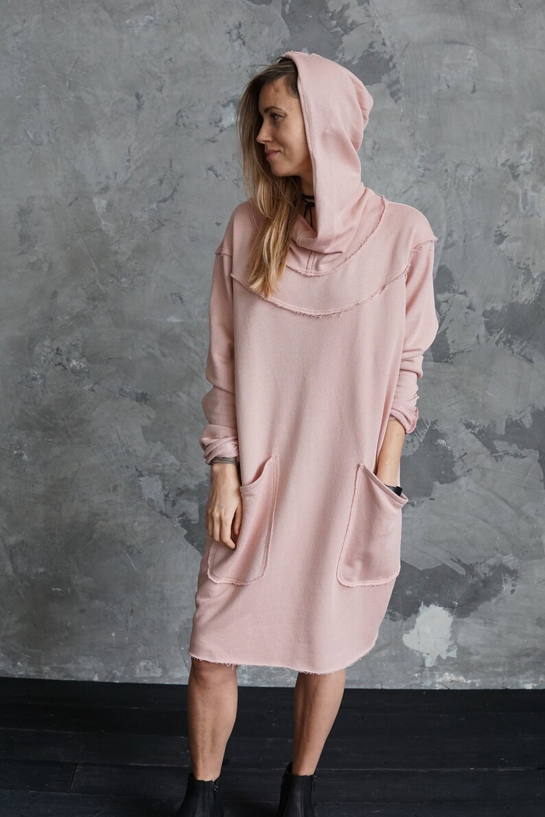 May include: A long, pink hooded sweatshirt dress with a cowl neck and two large pockets. The dress has a relaxed fit and a slightly distressed look.