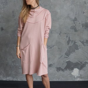 May include: A pink hooded sweatshirt dress with long sleeves and two large pockets. The dress has a distressed look with frayed edges.