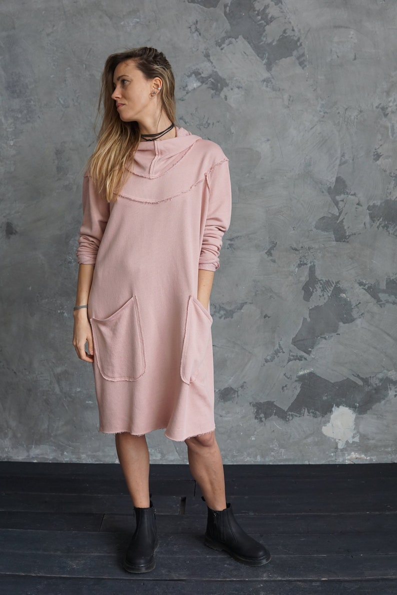 May include: A pink hooded sweatshirt dress with long sleeves and two large pockets. The dress has a distressed look with frayed edges.