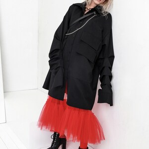 Op de afbeelding: Een zwart oversized shirt met een kettingdetail gedragen over een rode tule rok en zwarte laarzen met hak.