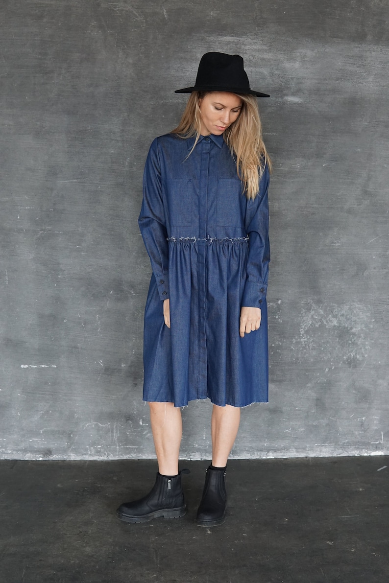 May include: A woman wearing a dark blue denim shirt dress with a gathered waist and long sleeves. She is wearing a black hat and black ankle boots.