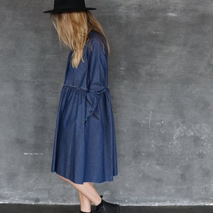 May include: A woman wearing a denim dress with a black hat and black ankle boots. The dress has a gathered waist and a raw hem.