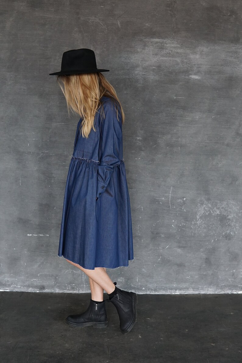 May include: A woman wearing a denim dress with a black hat and black ankle boots. The dress has a gathered waist and a raw hem.