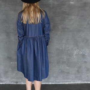 May include: A woman wearing a blue denim dress with a black hat and black ankle boots. The dress has a gathered waist and a slightly flared skirt.