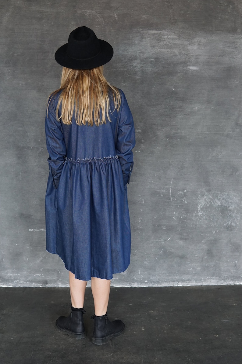 May include: A woman wearing a blue denim dress with a black hat and black ankle boots. The dress has a gathered waist and a slightly flared skirt.