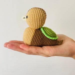 May include: A crocheted brown and tan duck with a green shell on its back. The duck is sitting on a hand.
