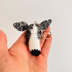May include: A crochet greyhound dog head keychain. The dog's head is white with black and grey speckled ears and a black nose. A silver keyring is attached. The keychain is handmade.