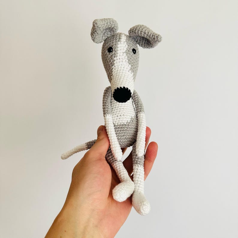 May include: A crocheted grey and white dog toy with black eyes and a black nose. The dog is sitting with its legs outstretched.