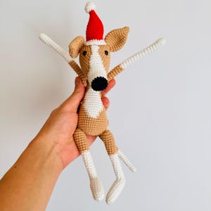 May include: A crocheted dog toy wearing a red Santa hat. The dog is brown and white with black eyes and a black nose. The toy is being held in a hand.