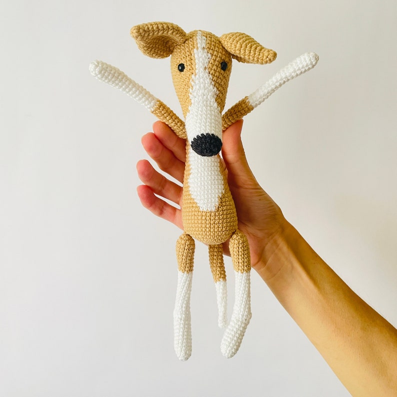 May include: A crocheted stuffed dog toy with brown and white fur. The dog has a black nose and white markings on its face. The toy is being held by a hand.