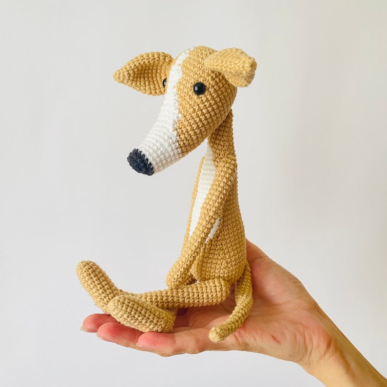 May include: A crocheted greyhound dog toy with white markings. The dog is sitting with its legs crossed and its tail curled around its body.