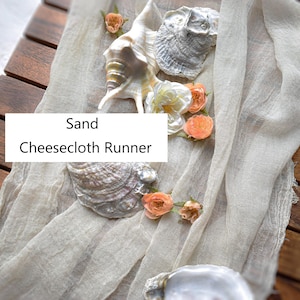 May include: A sand-colored cheesecloth runner draped over a wooden surface, decorated with various seashells and small peach-colored flowers. The text 'Sand Cheesecloth Runner' is overlaid on the center of the fabric runner.