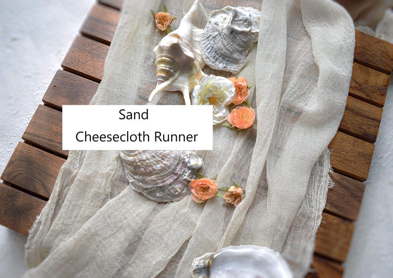 May include: A sand-colored cheesecloth runner draped over a wooden surface, decorated with various seashells and small peach-colored flowers. The text 'Sand Cheesecloth Runner' is overlaid on the center of the fabric runner.