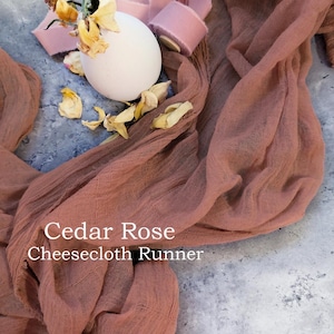 May include: A rustic brown cheesecloth table runner with the text "Cedar Rose Cheesecloth Runner" on it. The runner is folded and draped over a white surface.