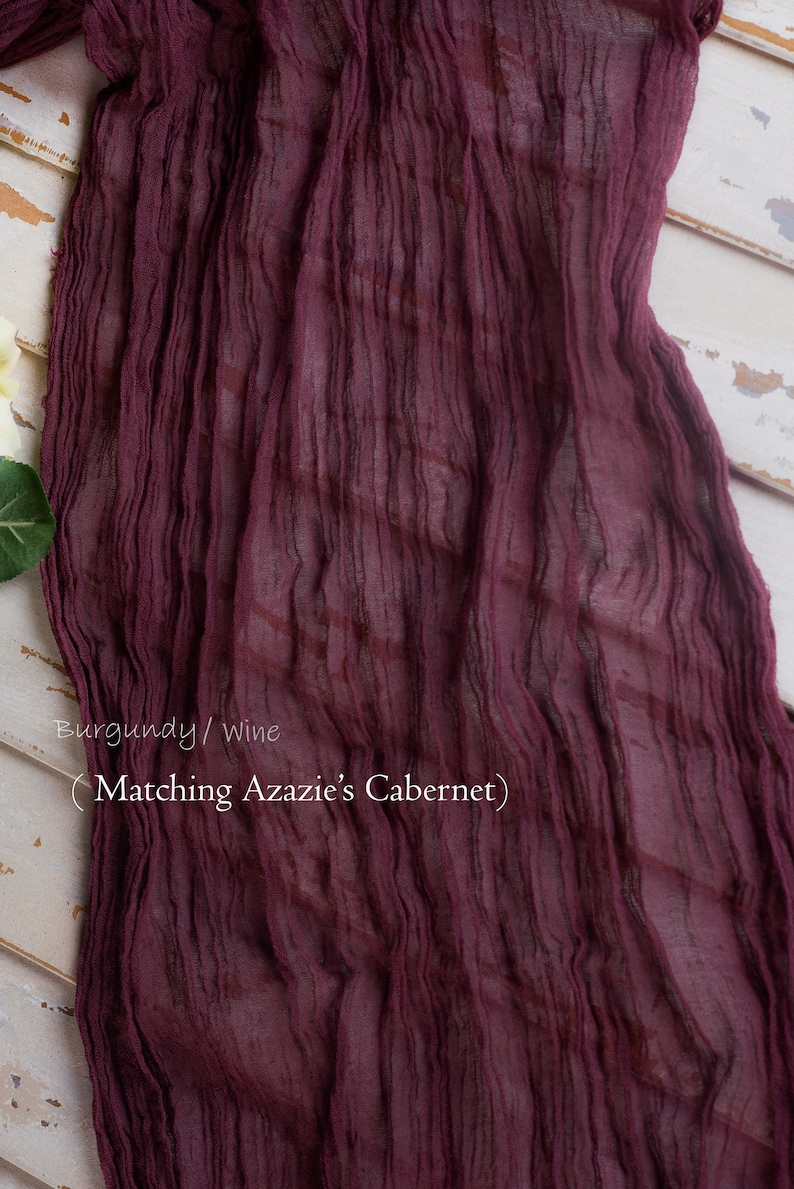 May include: Burgundy or wine colored sheer fabric with a crinkled texture. The fabric is draped over a white wooden surface. The text "Matching Azazie's Cabernet" is written on the fabric.