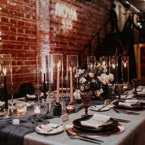 May include: A long table set for a formal dinner party with a grey tablecloth, black napkins, and gold chargers. The centerpiece is a large floral arrangement with pink roses and greenery. Tall glass cylinders with candles are placed along the table.