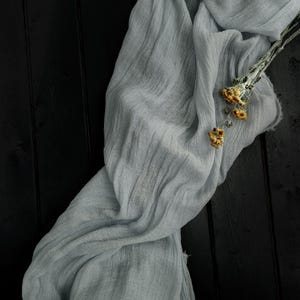 May include: A long, light gray, crinkled fabric runner with a sheer, gauzy texture. The fabric is draped in soft folds and features a small, dried sprig of yellow flowers resting on its surface, showcasing a delicate and lightweight appearance.