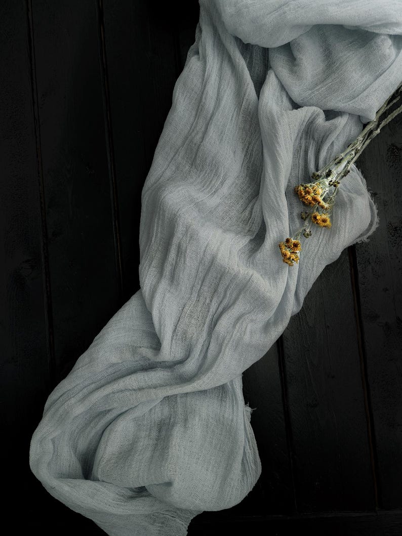 May include: A long, light gray, crinkled fabric runner with a sheer, gauzy texture. The fabric is draped in soft folds and features a small, dried sprig of yellow flowers resting on its surface, showcasing a delicate and lightweight appearance.