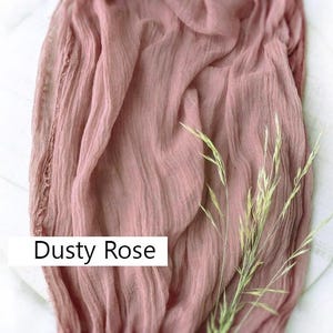 May include: Dusty rose colored sheer fabric with a delicate texture. The fabric is draped over a white surface with a sprig of dried grass.