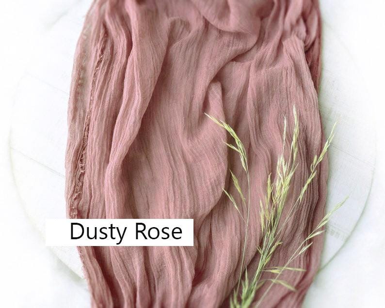 May include: Dusty rose colored sheer fabric with a delicate texture. The fabric is draped over a white surface with a sprig of dried grass.