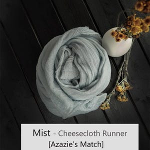 May include: A light blue, textured cheesecloth runner folded into a circular shape, accompanied by a small white vase containing dried yellow flowers. The text 'Mist - Cheesecloth Runner [Azazie's Match]' is printed in a white rectangular box.