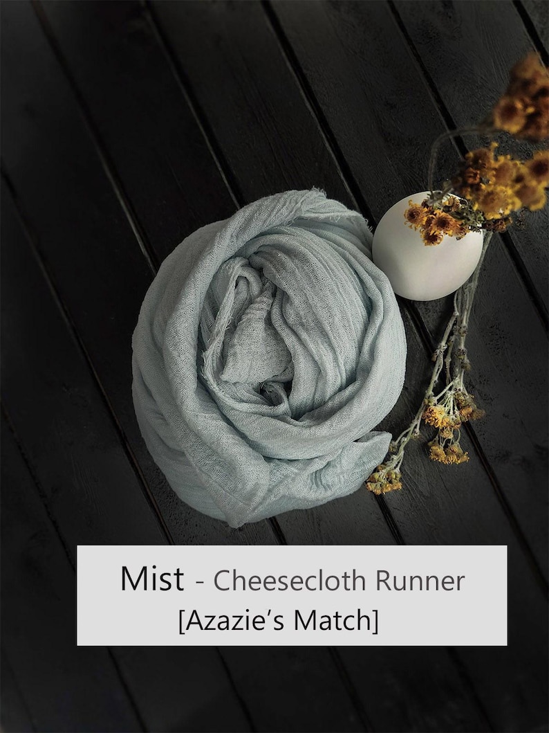 May include: A light blue, textured cheesecloth runner folded into a circular shape, accompanied by a small white vase containing dried yellow flowers. The text 'Mist - Cheesecloth Runner [Azazie's Match]' is printed in a white rectangular box.