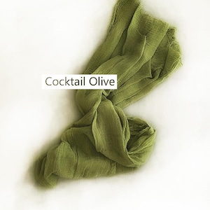 May include: A long, rectangular piece of olive green fabric with a crinkled, textured surface. The fabric is draped in a loose, flowing manner. A white rectangular label with the black text Cocktail Olive is positioned over the fabric.