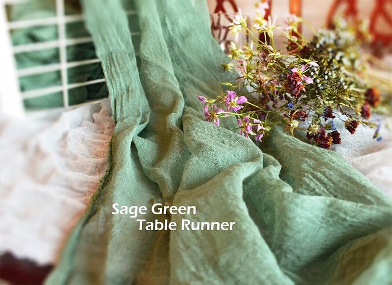May include: A sage green table runner with a crinkled, textured fabric surface. The text "Sage Green Table Runner" is printed in white on the fabric. A small bouquet of dried wildflowers with pink, purple, and yellow petals rests on the runner.