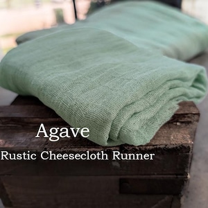 May include: A folded sage green cheesecloth runner with a loose, gauzy weave texture. The fabric is draped over a dark brown wooden crate. The text Agave Rustic Cheesecloth Runner is printed in white on the front of the wooden crate surface.