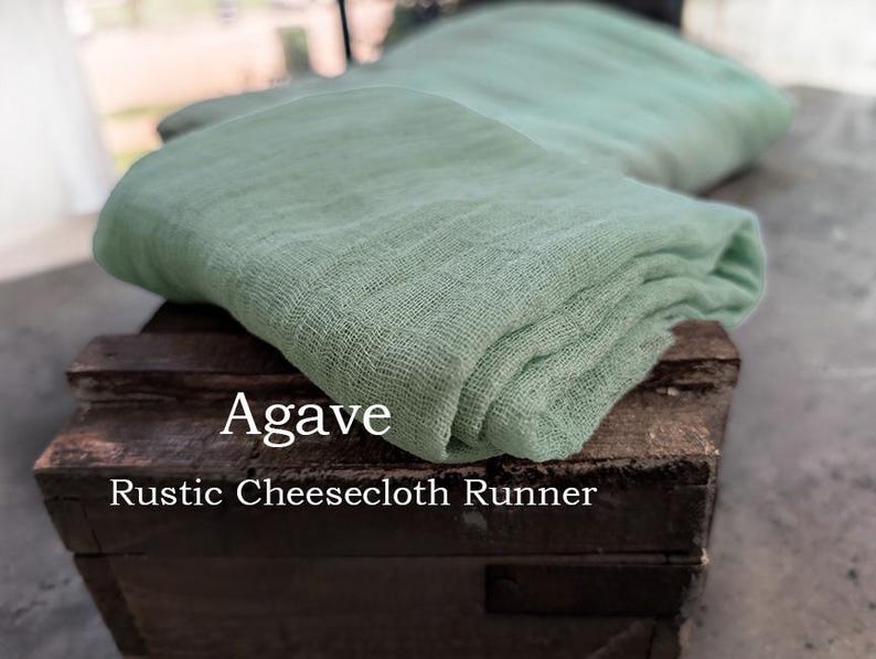 May include: A folded sage green cheesecloth runner with a loose, gauzy weave texture. The fabric is draped over a dark brown wooden crate. The text Agave Rustic Cheesecloth Runner is printed in white on the front of the wooden crate surface.