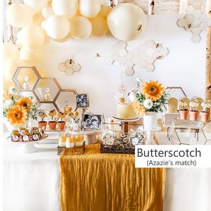 May include: A party dessert table featuring a white tablecloth with a mustard yellow runner, decorated with cream-colored balloons, honeycomb-shaped wall accents, sunflowers, small potted plants, glass jars, and a cake with the text Butterscotch (Azazie's match).