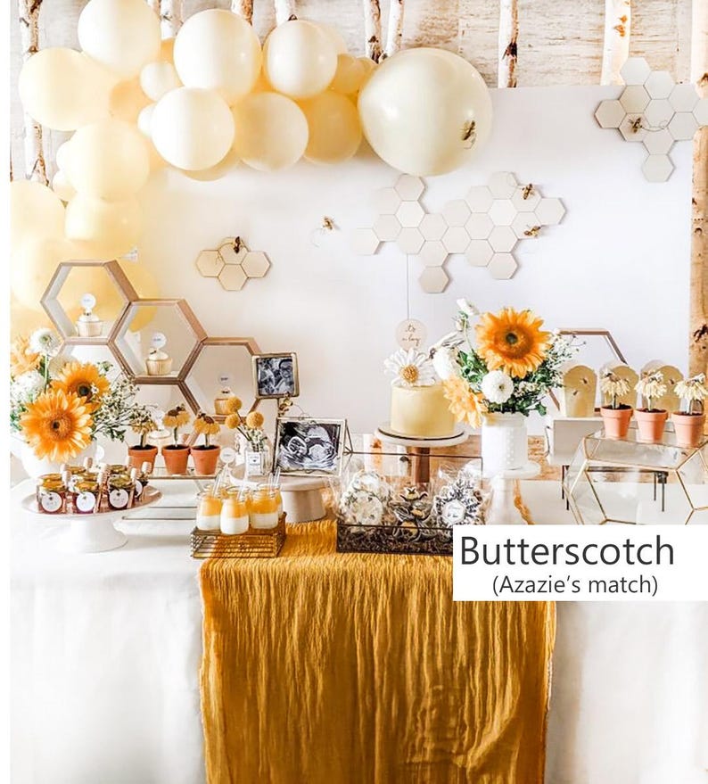May include: A party dessert table featuring a white tablecloth with a mustard yellow runner, decorated with cream-colored balloons, honeycomb-shaped wall accents, sunflowers, small potted plants, glass jars, and a cake with the text Butterscotch (Azazie's match).