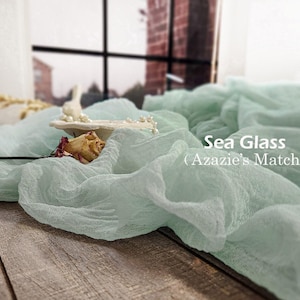 May include: A sea glass colored, textured cheesecloth fabric runner draped in folds. The text 'Sea Glass (Azazie's Match)' is printed in white on the right side. A small white decorative dish with pearls and a dried rosebud rests on the fabric.