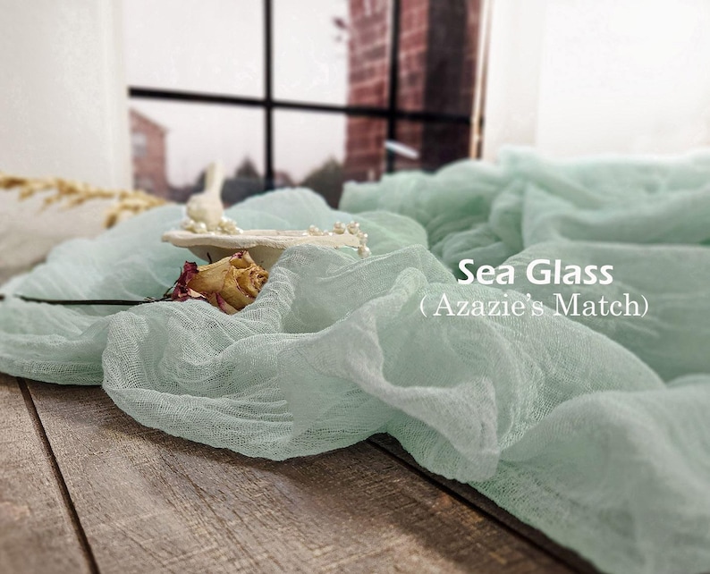 May include: A sea glass colored, textured cheesecloth fabric runner draped in folds. The text 'Sea Glass (Azazie's Match)' is printed in white on the right side. A small white decorative dish with pearls and a dried rosebud rests on the fabric.